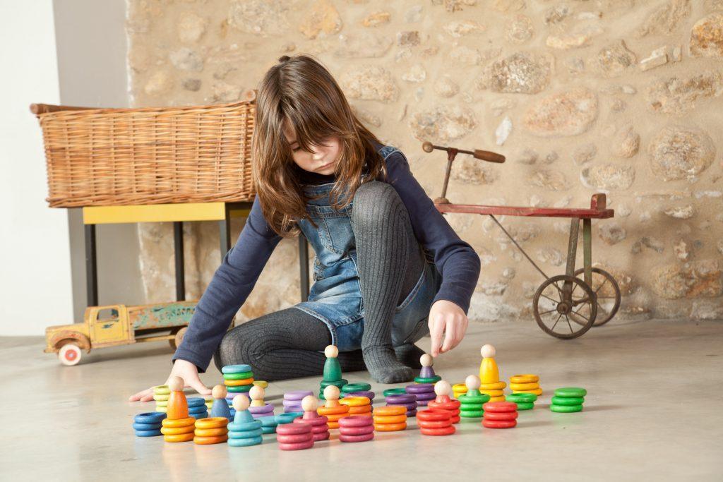 A young girl sits on the floor playing with colorful Grapat Nins Carla, focused on stacking them to enhance her hand-eye coordination, against a backdrop featuring a stone wall and a vintage toy wagon.