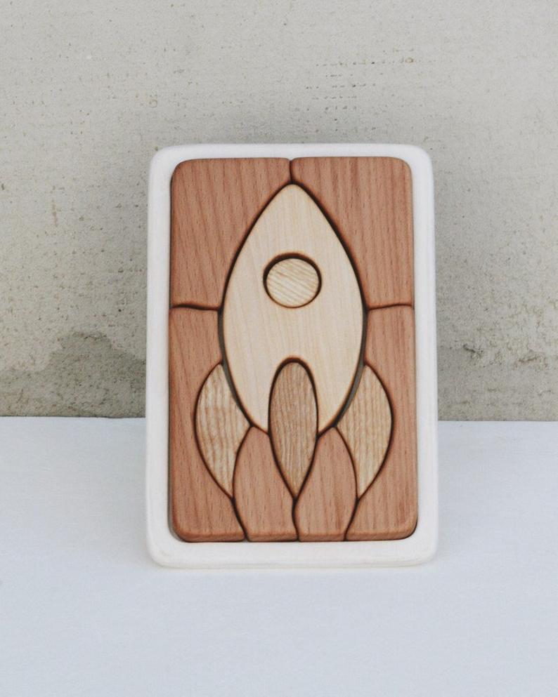 A Handmade Wooden Puzzle, handmade in Russia, features a stylized rocket ship design. The puzzle pieces vary in shades of cherry wood, fitting neatly within a white frame. The background is a plain light grey surface.