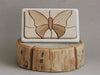 A Handmade Wooden Puzzle, handmade in Russia and carved with a butterfly design, features varying shades of Cherry wood. It is displayed upright on a rustic, cut tree stump base against a plain, light gray background. The piece is beautifully finished with linseed oil to enhance its natural beauty.