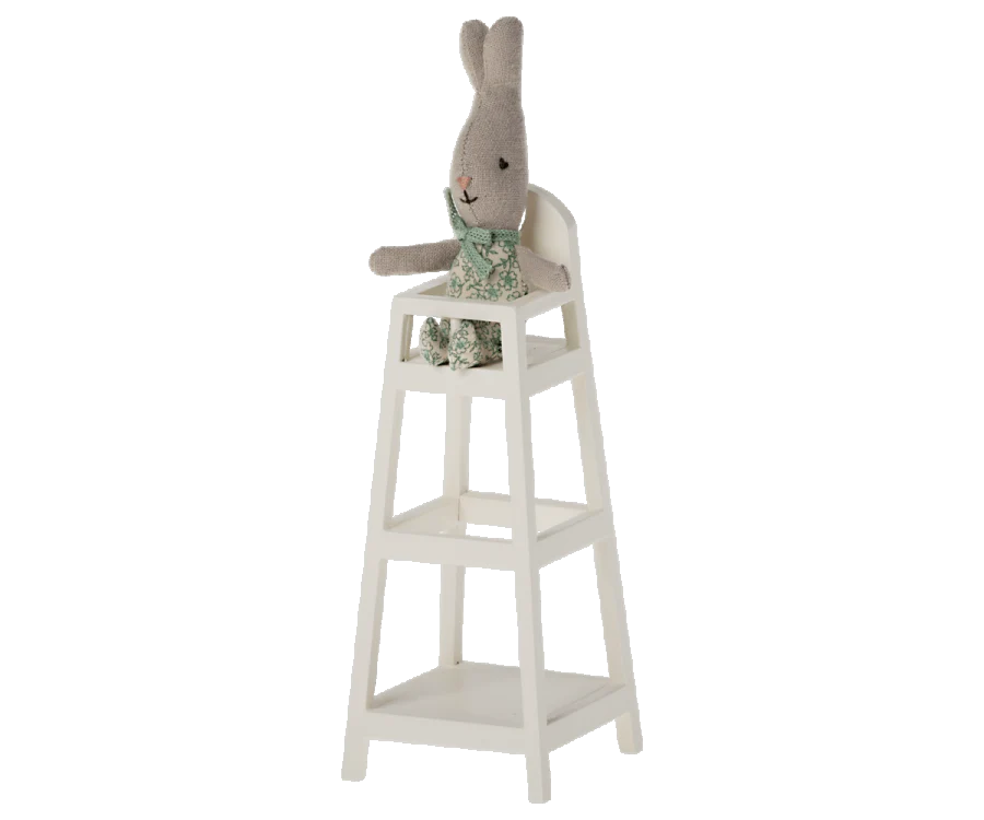 A gray stuffed rabbit wearing green floral overalls sits on the top seat of a Maileg My High Chair. The baby high chair features three tiers below the seat. The rabbit’s head has long ears and a stitched smile, adding to the overall minimalist, whimsical feel.