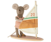 A Maileg Little Sister Surfer Mouse wearing a peach-colored outfit stands on a toy windsurf board, holding onto the white frame attached to a fabric sail. The sail, marked with the number "20," features horizontal green, orange, and yellow stripes. Elastic straps secure its magnetic hands to the frame for added stability.