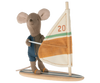 A Maileg Little Brother Surfer Mouse, dressed in a blue wetsuit, is holding onto the sail of a small windsurfing board using its magnetic hands. The sail is white with orange and green stripes, and the number 20 is printed on it. The mouse stands on a wooden base that resembles a surfboard.