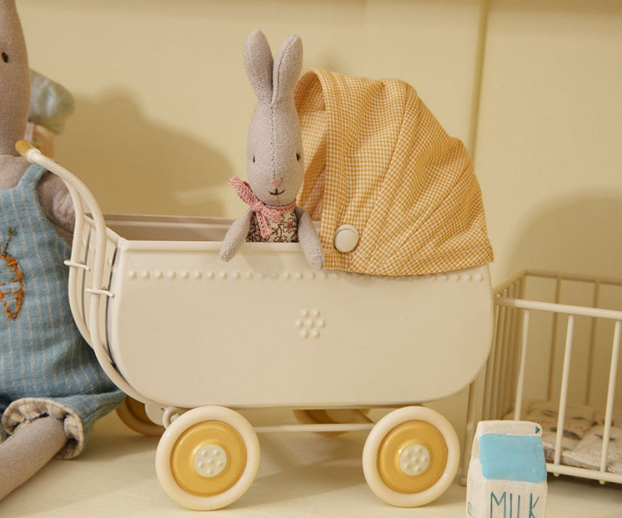 A Maileg Micro Pram with a toy rabbit inside, covered by a checkered cloth, beside a toy bear and a blue block with "milk" written on it.