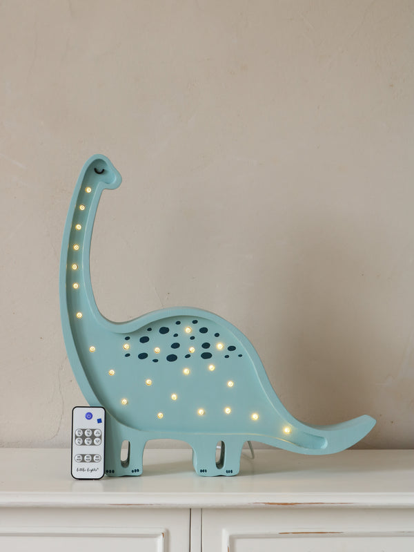 A blue Little Lights Dinosaur Lamp stands proudly on a white surface against a beige wall, perfect for nursery decor. Crafted from handmade natural pinewood, it features small glowing lights and comes with a remote control. The dinosaur boasts a long neck, tail, and cut-out patterns for the lights.