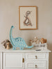 A white dresser topped with a Little Lights Dinosaur Lamp, a crocheted bunny toy, and a white wire basket holding wooden toys sits as the centerpiece of this charming nursery decor. Above it hangs a framed illustration of a dinosaur, set against a textured light beige wall. The setting exudes warmth and whimsy.