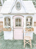 A quaint children's playhouse painted in white with pink doors and trim, featuring a round window on the upper facade and flower boxes under side windows, with a Toddler Rattan Bow Chair (Set of Two) in front.