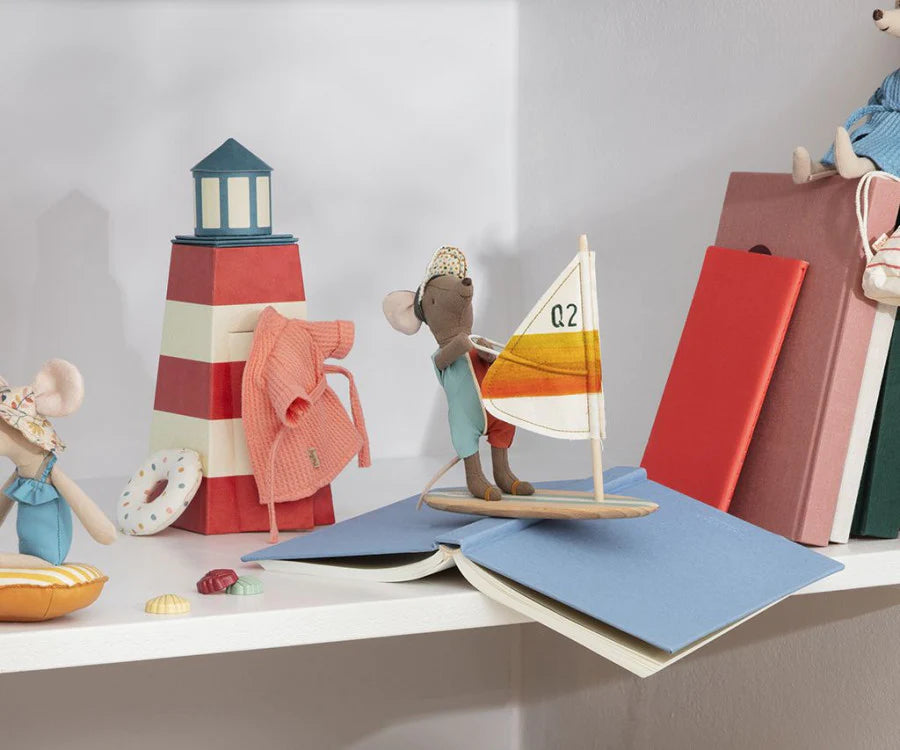 A whimsical scene featuring a small toy mouse holding a sailboat on an open blue book. Surrounding it are colorful books, a miniature striped lighthouse, shells, and a Maileg Surfer Big Brother Mouse dressed in blue sitting nearby with a wooden surfboard. The background is a light-colored wall.