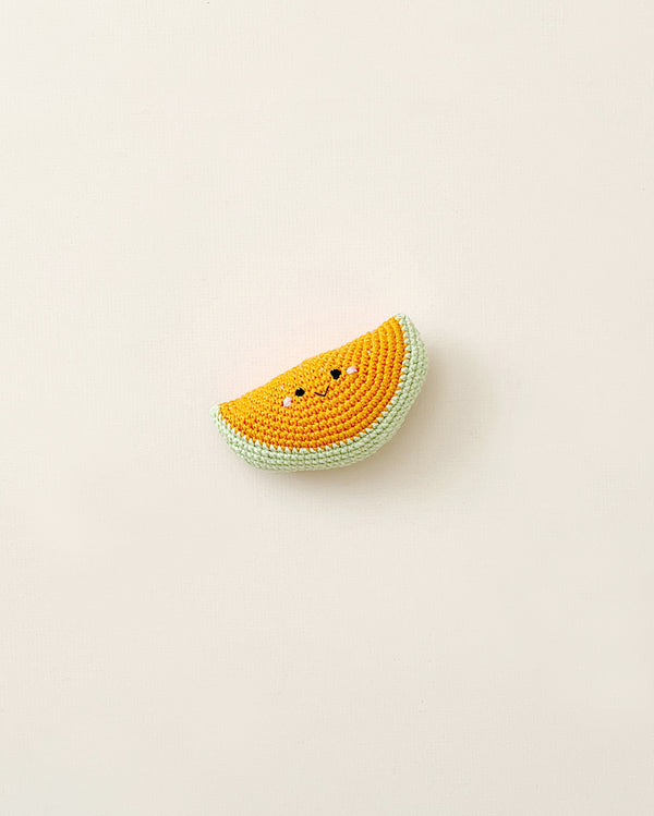 A Handmade Melon Rattle resembling a slice of melon with a smiling face, crocheted from cotton yarn and displayed on a plain, light-colored background.