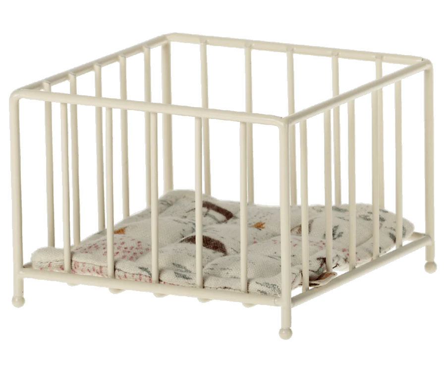 A small, white metal playpen with bars on all sides. Inside the Maileg Playpen, there is a cushioned pad with a patterned fabric featuring various illustrations and a cute cushion that's perfect for your My-sized Maileg.