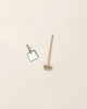 A minimalist image featuring a bamboo toothbrush lying horizontally and a small mirror with a gold frame set at an angle, alongside a Maileg Miniature Broom Set on a light pink background.