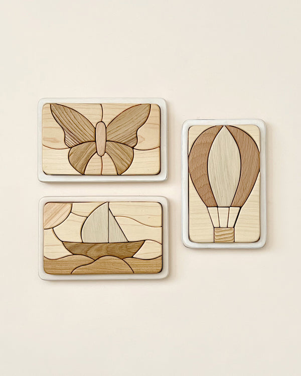 Three Handmade Wooden Puzzle pieces with puzzle-like designs, handmade in Russia. The top-left piece features a butterfly, the top-right shows a hot air balloon, and the bottom-left depicts a sailboat. Each piece combines different shades of light and dark wood, finished with linseed oil for a polished look.