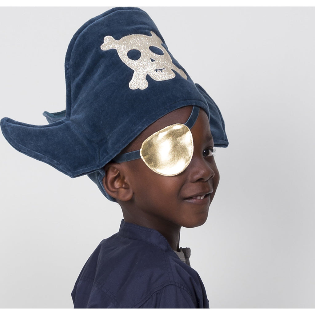 A young boy wearing a Meri Meri Pirate Costume with a large blue hat adorned with a skull and crossbones, and a shiny gold eye patch, smiles at the camera during a pirate party.