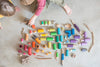 Two children sitting by an arrangement of colorful craft materials including threads, Grapat Lola figures, beads, and natural items like twigs and stones on a light grey background.