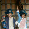 Two children engaged in imaginative play, dressed in Meri Meri Pirate Costumes, one holding a sword and lantern, smiling and posing in front of an old wooden door.