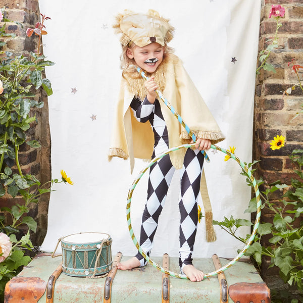 A joyful child in a Meri Meri Lion Costume headdress and harlequin costume plays with a hula hoop, amid musical drums and greenery, displaying playful energy.