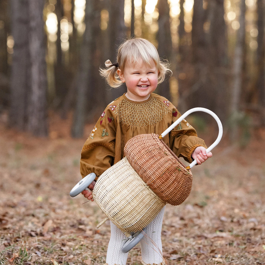 A joyful toddler with blonde hair, wearing a brown blouse and white pants, laughs while holding an Olli Ella Rattan Mushroom Luggy - Natural basket in a forest during autumn.