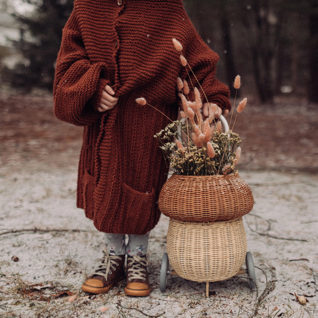 A person in a chunky knit maroon cardigan stands on a snowy path, partially obscuring a whimsical Olli Ella Rattan Mushroom Luggy - Natural filled with dried flowers and grasses.