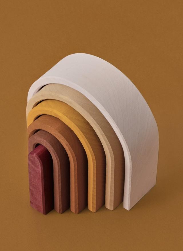 A stack of Raduga Grez Handmade Wooden Oval Rainbow Stackers crafted from linden wood in varying sizes and shades from cream to dark brown, arranged in a semi-circular formation against a plain beige background.