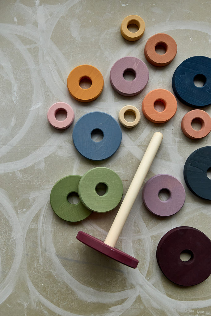 Assorted colorful wooden rings and a stick on a textured gray background, part of a Raduga Grez | Handmade Large Pyramid Tower Stacker - Colorful designed to help develop hand-eye coordination and fine motor skills.