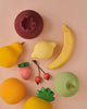 A collection of Raduga Grez Handmade Painted Wooden Fruits arranged on a light pink background. There are bananas, apples, a strawberry, cherries, a pear, and a lemon, creating a playful and artistic display.