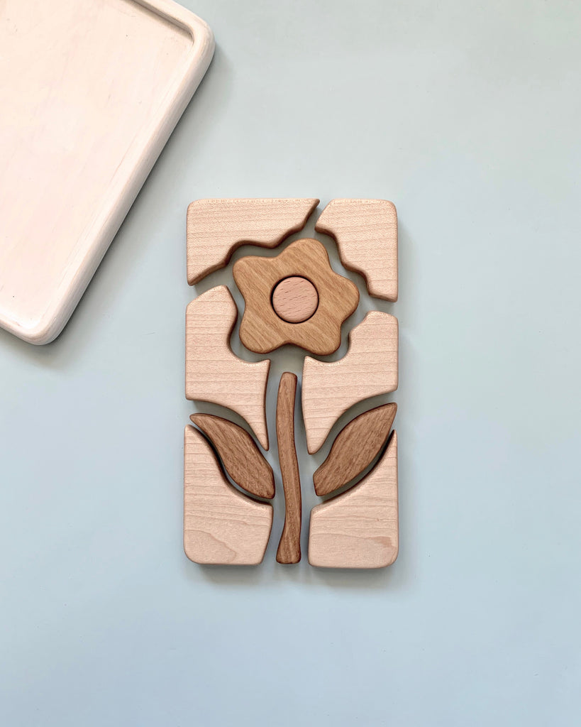 A Handmade Wooden Puzzle of a flower with a stem and leaves is assembled on a light blue surface. Crafted from cherry wood and treated with linseed oil, the pieces fit neatly together to form a complete flower image. A rectangular wooden tray is partially visible in the top left corner.