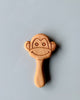 A Handmade Wooden Teether shaped like a monkey's face with a smiling expression is positioned against a plain grey background. The handle extends downwards, and the rattle, crafted from cherry wood with minimalistic detail, ensures it is a non-toxic baby teether.