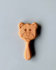 A Handmade Wooden Teether shaped like a bear's face with engraved eyes and a smiling mouth. The non-toxic teether handle is smooth and extends below the bear's head, crafted entirely from cherry wood. The background is a plain, light gray surface.