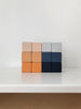 A minimalistic arrangement of twelve wooden blocks from Sabo Concept is displayed on a white surface. The Mini Wooden Blocks - Desert Night handmade stacking toy is organized in a 3x4 grid, featuring various shades of beige, blue, and orange painted with non-toxic paint. The background is plain white.