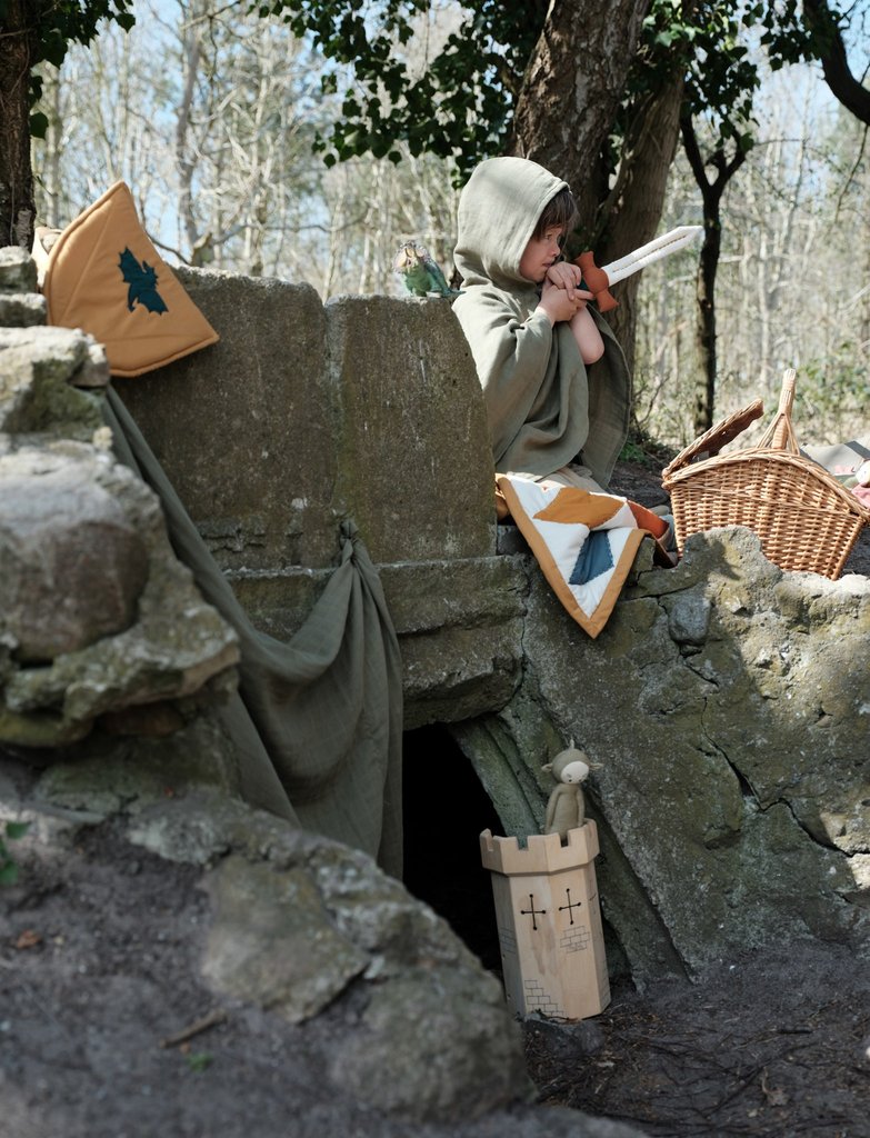 A picture of a boy playing out in nature dressed as a knight, holding a fabric sword.