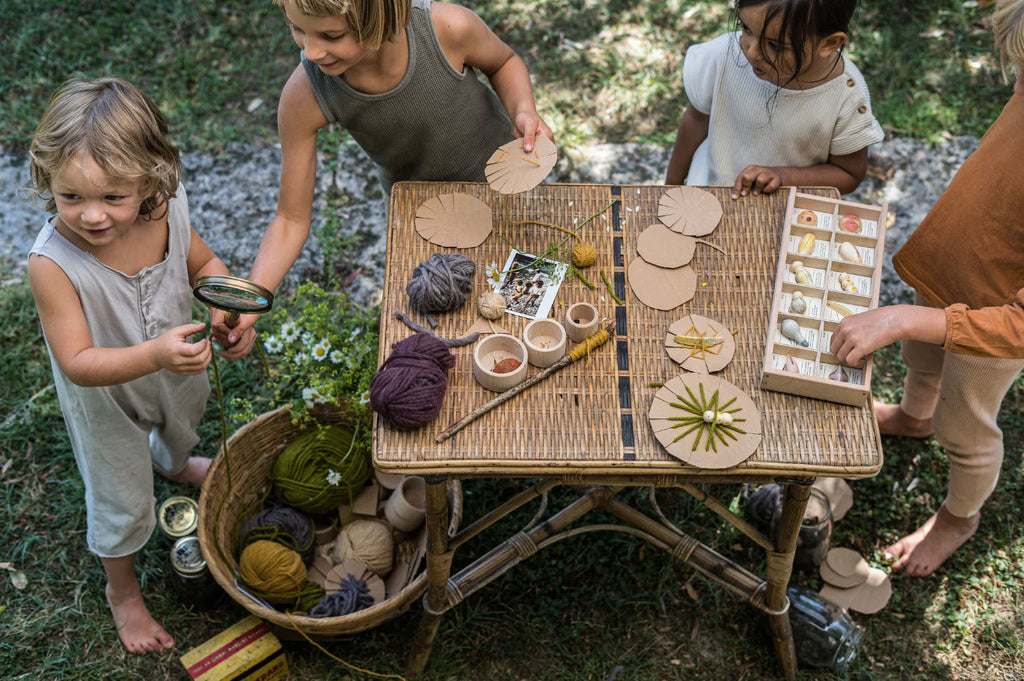 Children engaged in a nature craft activity outdoors, using creatures from the fauna realm as part of the Grapat Wild Exploration Play Set on a wicker table, under the shade of trees.