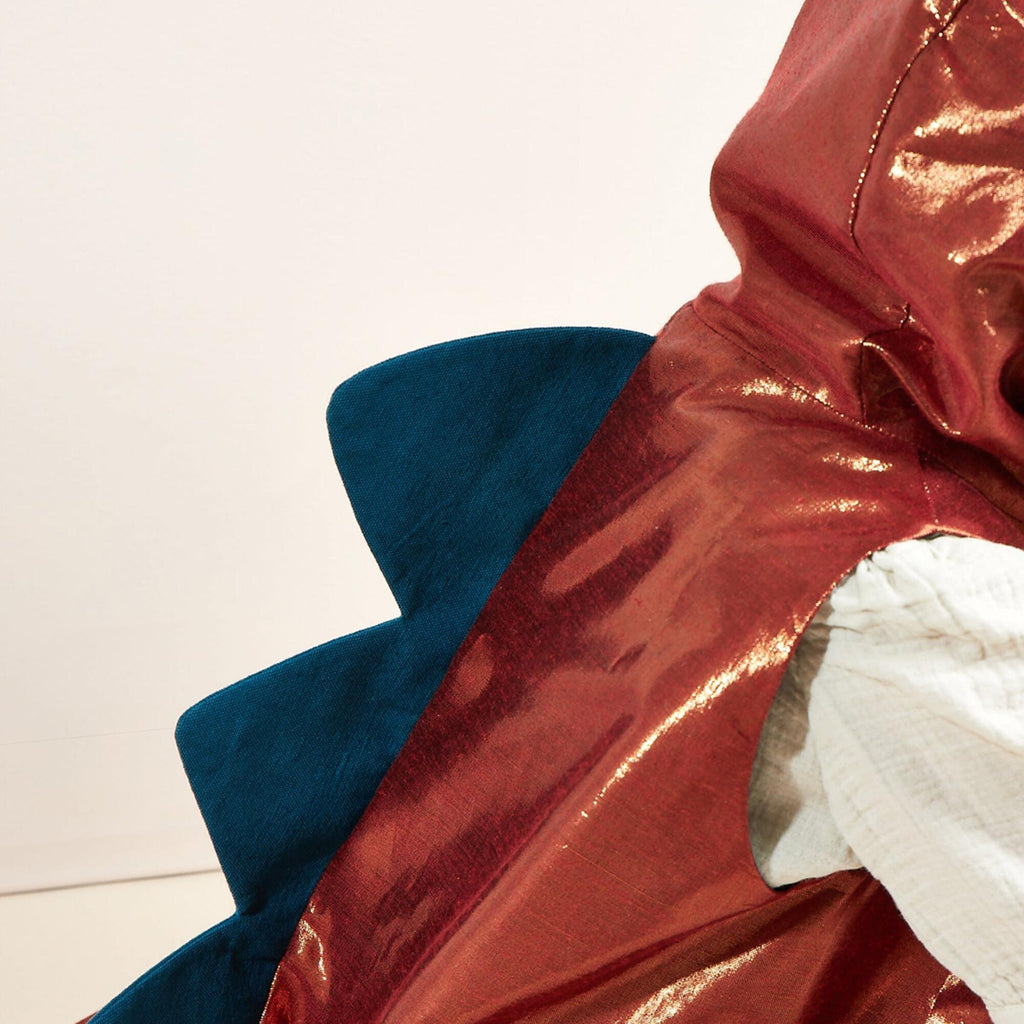 A close-up of a shiny red Meri Meri Dinosaur Costume - Final Sale with blue spikes along the back. The costume is worn by someone, with part of a white sleeve visible on the right side. The background is plain and light-colored, making this kids costume stand out marvelously.